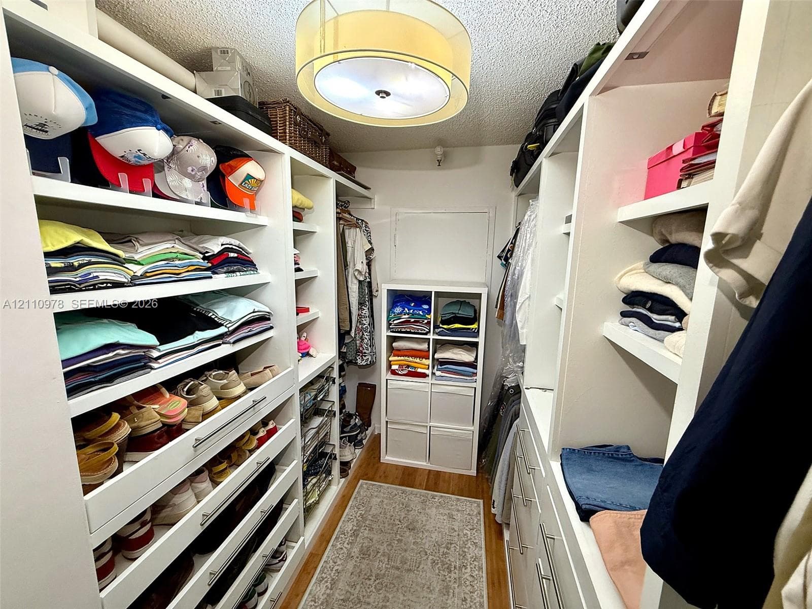 Custom walk-in closet featuring extensive white built-in shelving, integrated storage drawers with polished hardware, wood flooring, and a drum-style ceiling light fixture