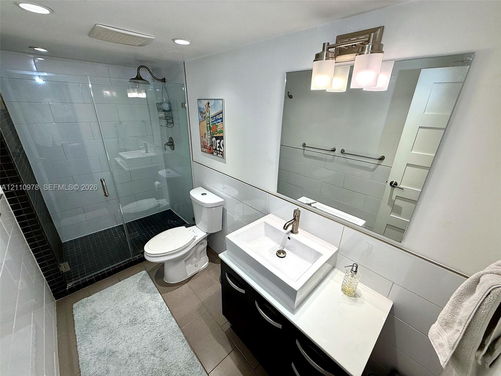 Walk-in shower with a glass enclosure, rectangular vessel sink, dark-finish vanity cabinetry, and wall-mounted lighting