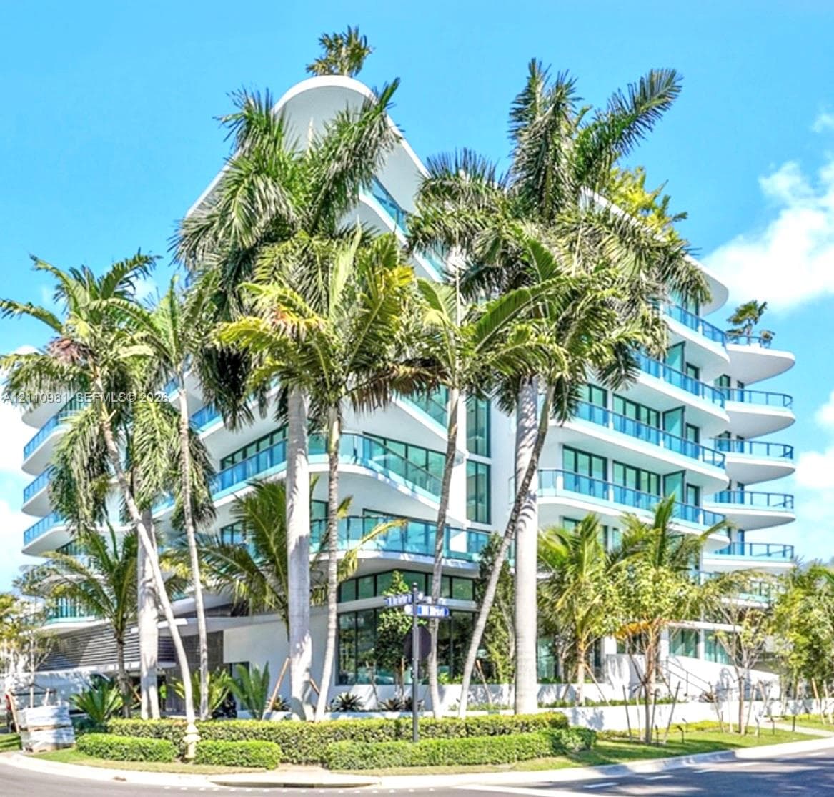 Modern multi-story structure featuring curved cantilevered balconies, floor-to-ceiling glass paneling, white exterior finish, and surrounding mature palm landscaping