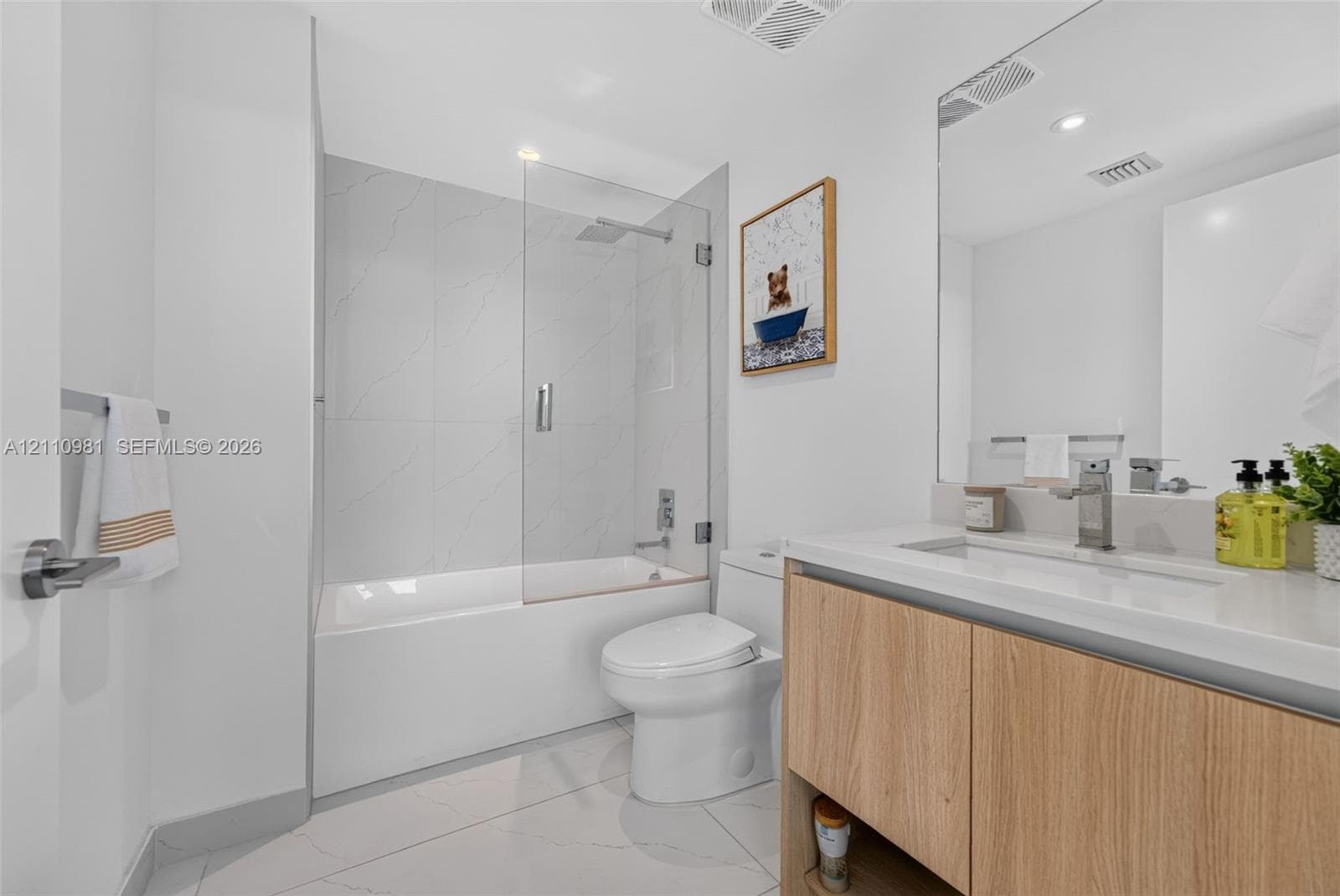 Bathroom featuring a wood-finish vanity with a stone-surface countertop, an integrated sink with a chrome faucet, large-format tile flooring, and a shower-tub combination enclosed by a frameless glass panel with chrome fixtures