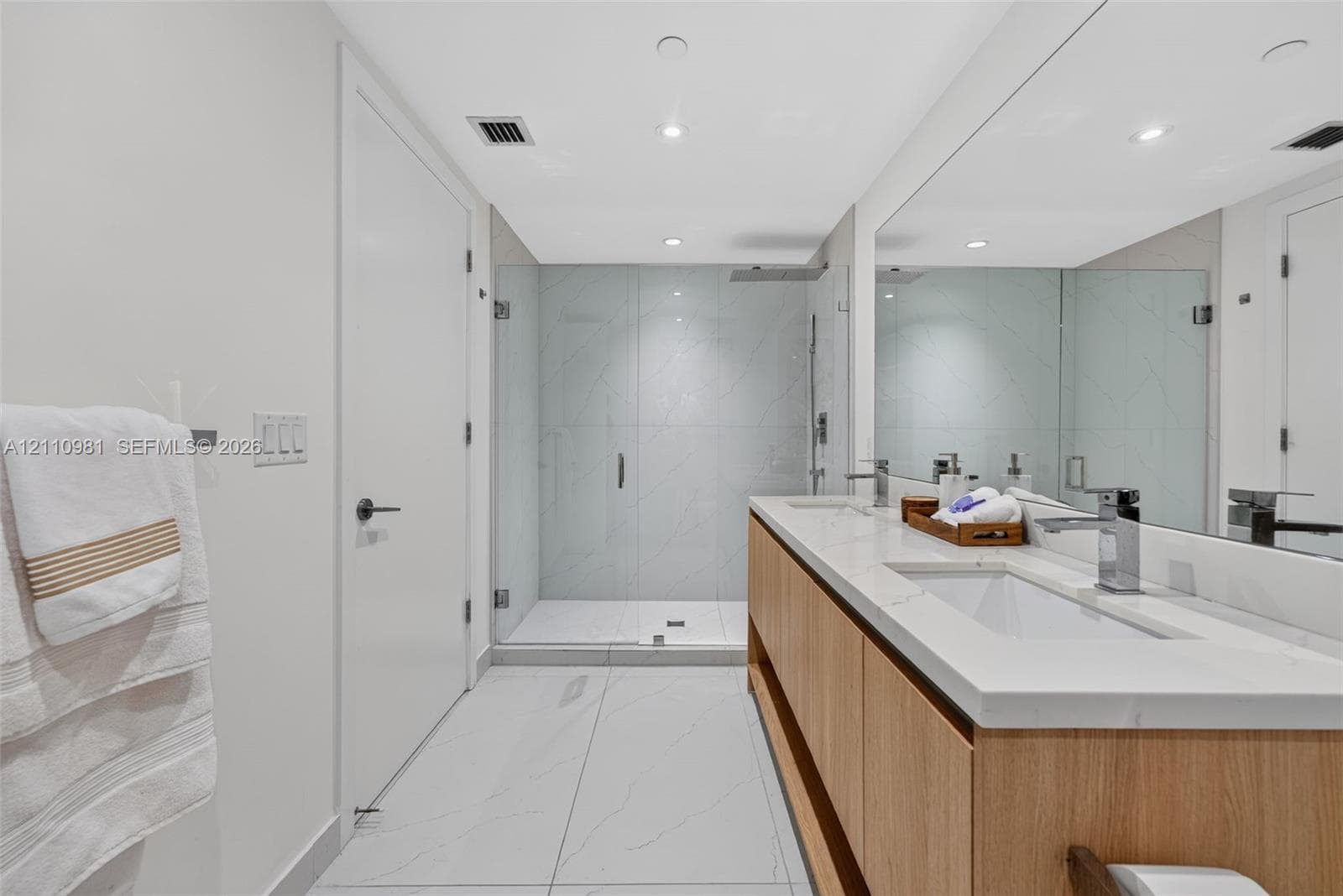 Dual-sink vanity with light wood cabinetry, quartz countertops, and polished chrome faucets, complemented by a glass-enclosed shower with large-format stone-patterned tile flooring and wall surfacing