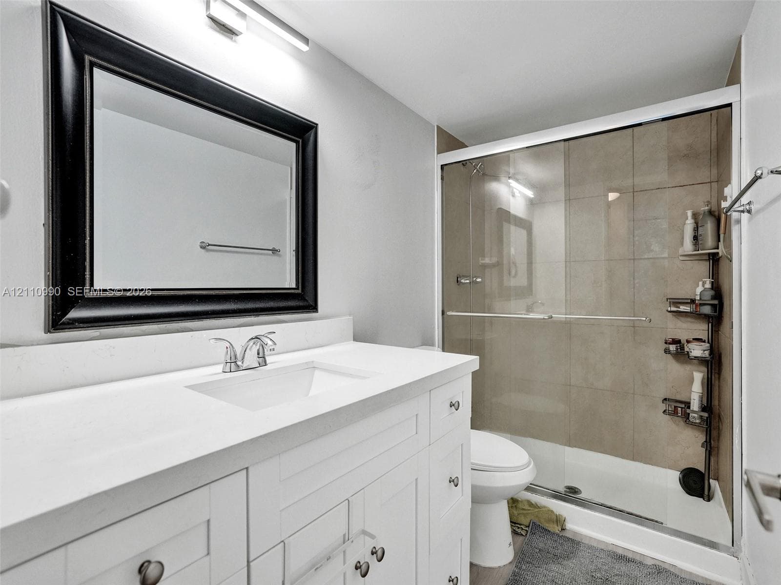White shaker-style vanity with a solid surface countertop, rectangular integrated sink, chrome faucet, framed mirror, and tiled shower enclosure with a glass sliding door