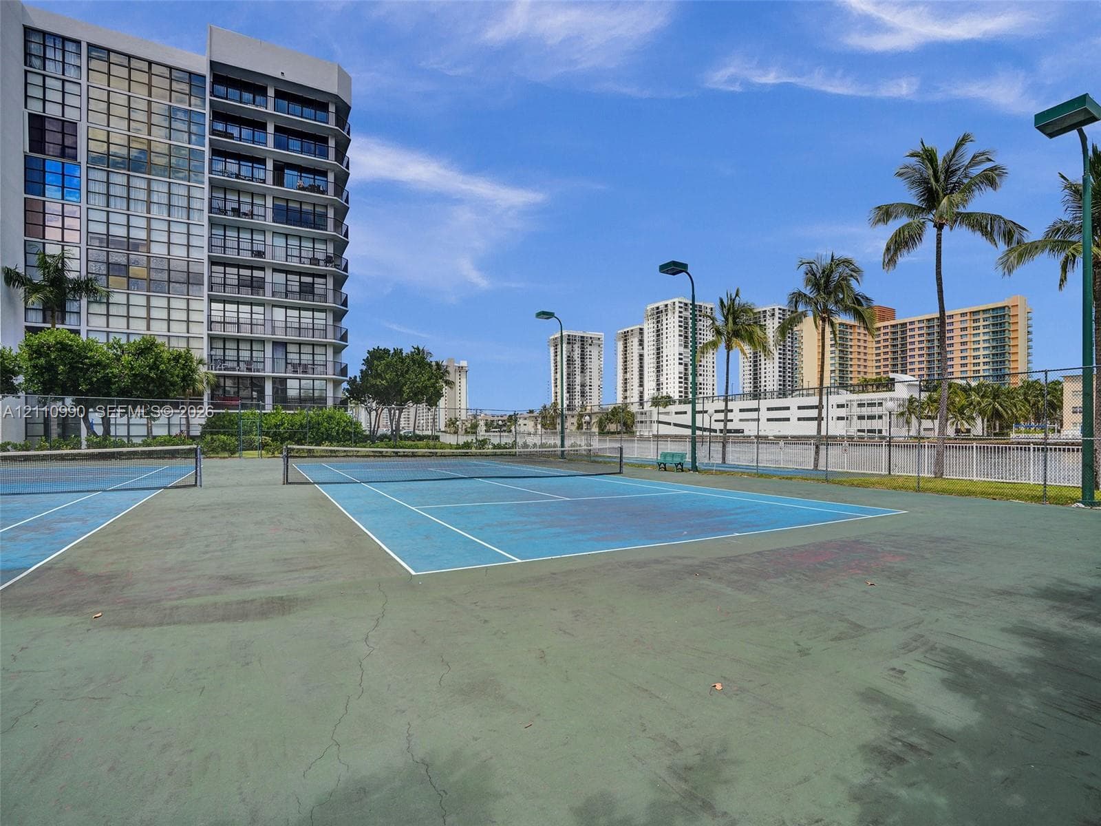 Hard-court tennis facility featuring perimeter chain-link fencing, dual court lighting, and mature palm trees