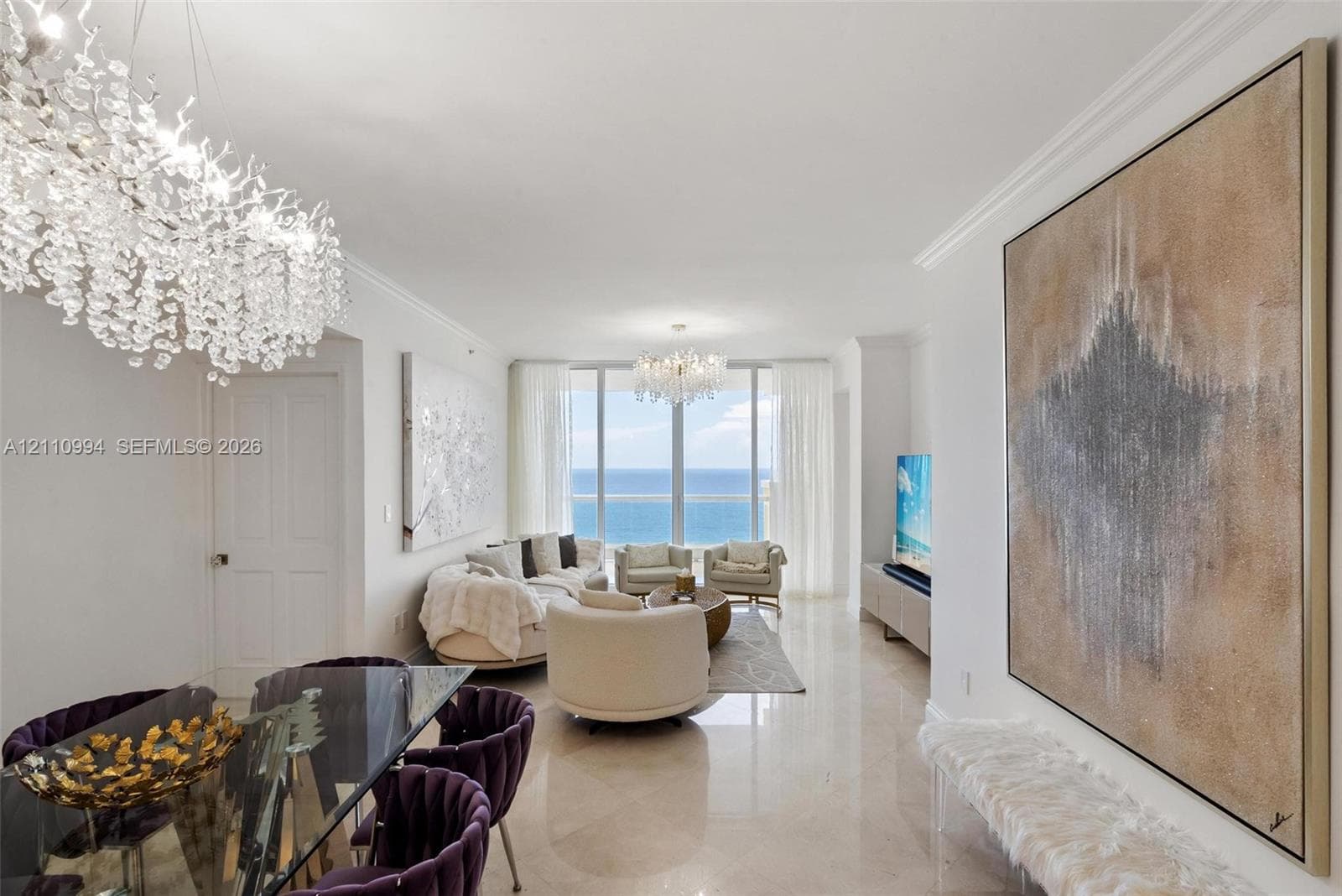 Expansive living area featuring floor-to-ceiling windows with ocean scenery, polished stone flooring, recessed crown molding, and dual crystal chandeliers