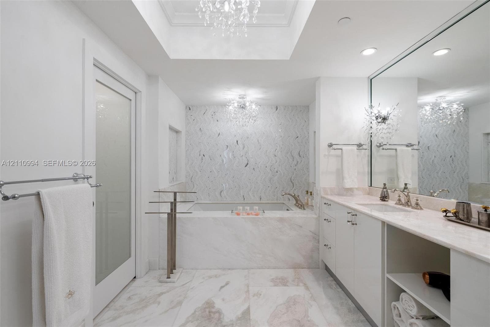 Soaking tub with stone surround, custom tiled accent wall, skylight with recessed tray ceiling, dual crystal chandeliers, and white cabinetry with stone countertop
