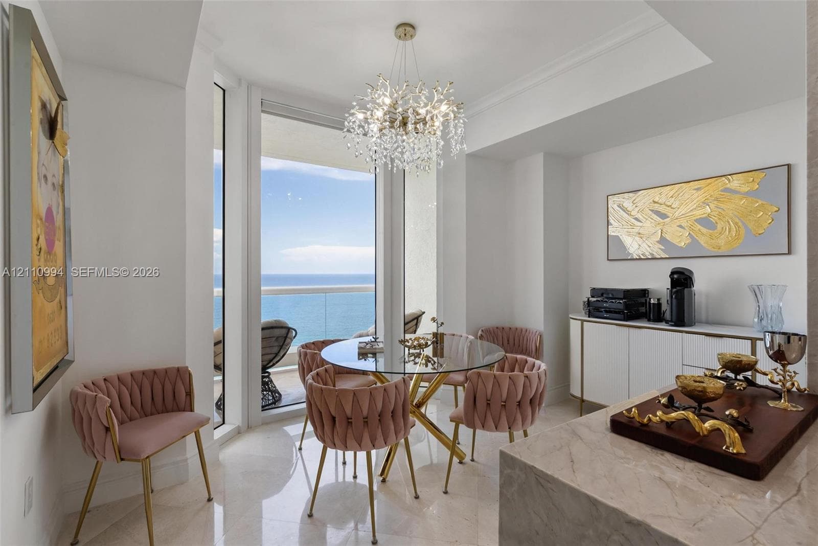 Dining area featuring large-format polished stone flooring, floor-to-ceiling windows with ocean horizon, a multi-tiered crystal chandelier, and built-in white cabinetry with gold-tone hardware