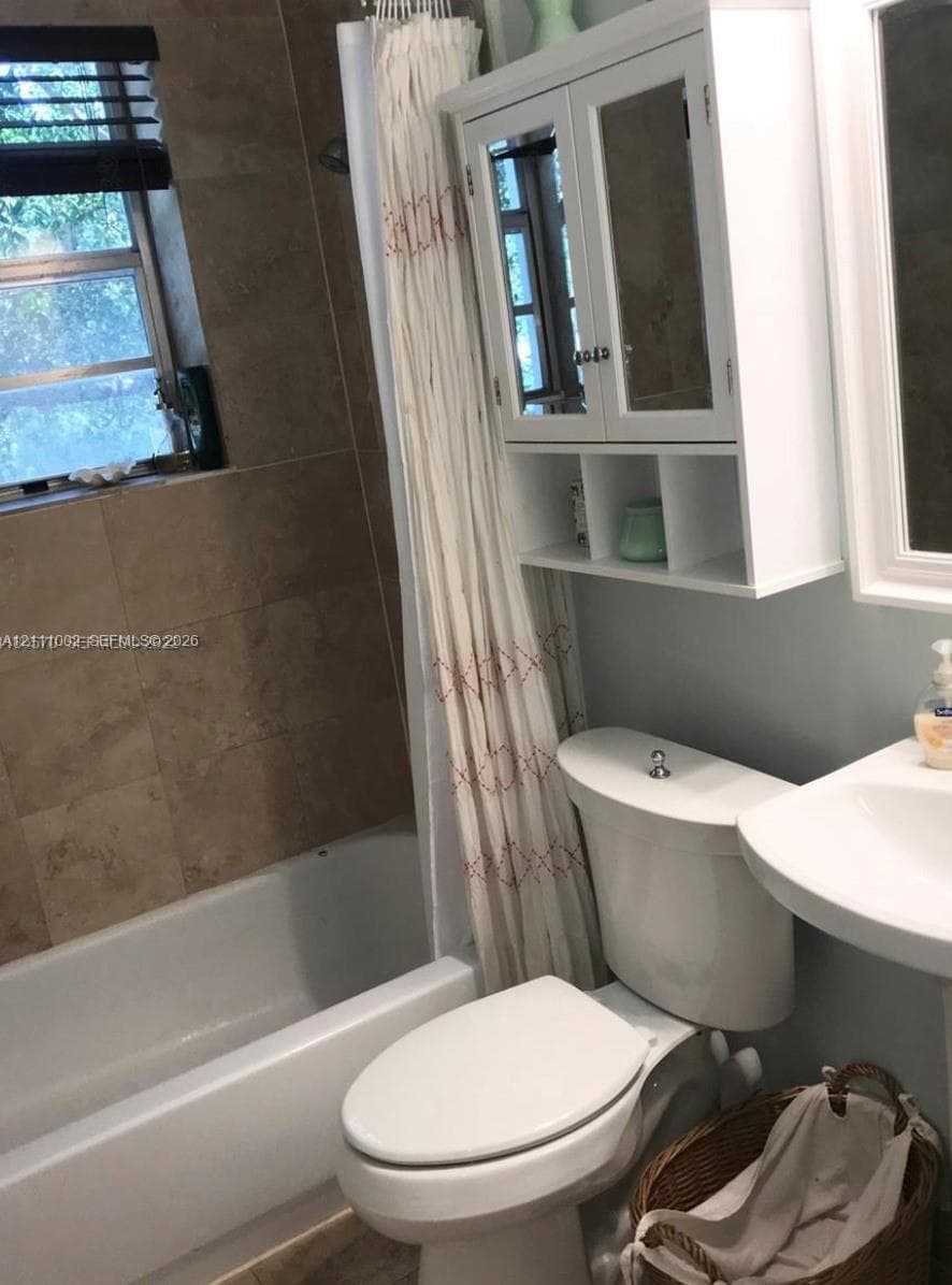 Bathroom featuring a white pedestal sink, a built-in white wall cabinet with glass-panel doors, a standard white toilet, and a shower-tub combination finished with neutral wall tiling