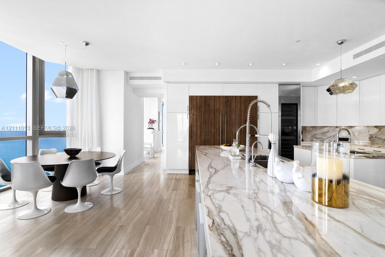 Contemporary kitchen featuring a large stone island with an integrated sink, high-gloss cabinetry, wood-finish flooring, and floor-to-ceiling windows