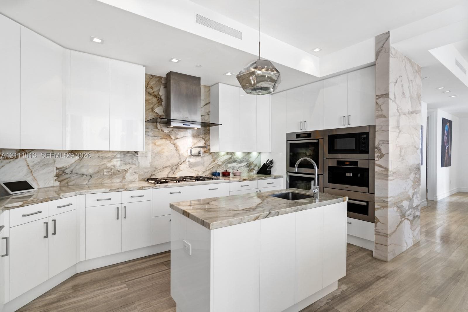 Contemporary kitchen featuring high-gloss white cabinetry, expansive stone slab countertops and backsplash, integrated stainless steel double wall ovens, wood-finish flooring, and a central island with an undermount sink