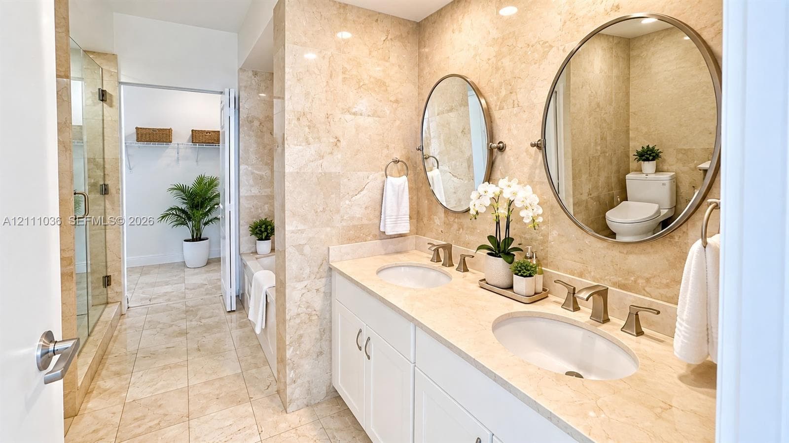 Double vanity featuring white cabinetry, stone-finish countertops, dual oval mirrors, and wall-mounted metallic fixtures, accented by floor-to-ceiling stone wall tiling and a glass-enclosed shower
