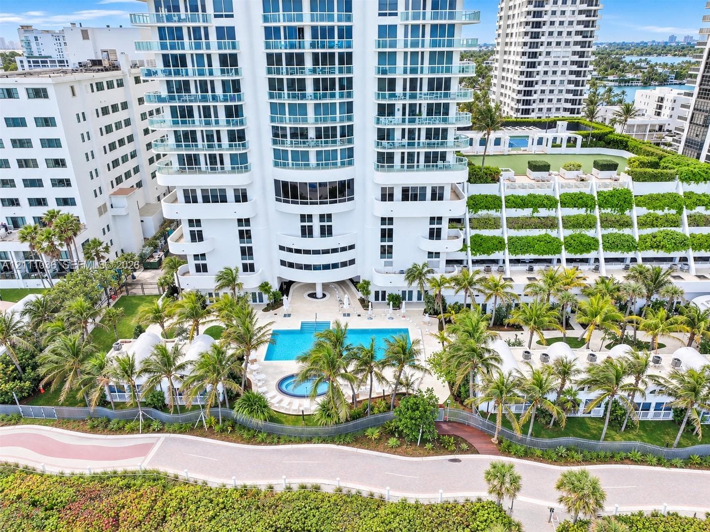 High-rise residential exterior featuring expansive glass balconies, a ground-level swimming pool, a separate spa, landscaped grounds with mature palm trees, and multi-level terraced gardens
