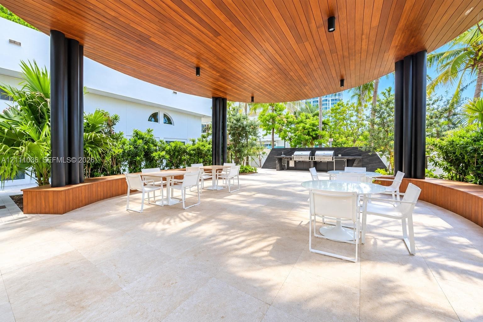 Covered outdoor terrace featuring a wood-paneled ceiling, stone tile flooring, structural cylindrical columns, and a built-in outdoor kitchen station with stainless steel grills
