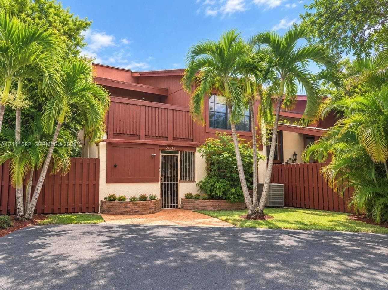 Multi-level exterior featuring a private upper-level balcony, decorative brick masonry planters, paved entry walkway, and established palm landscaping enclosed by wood privacy fencing