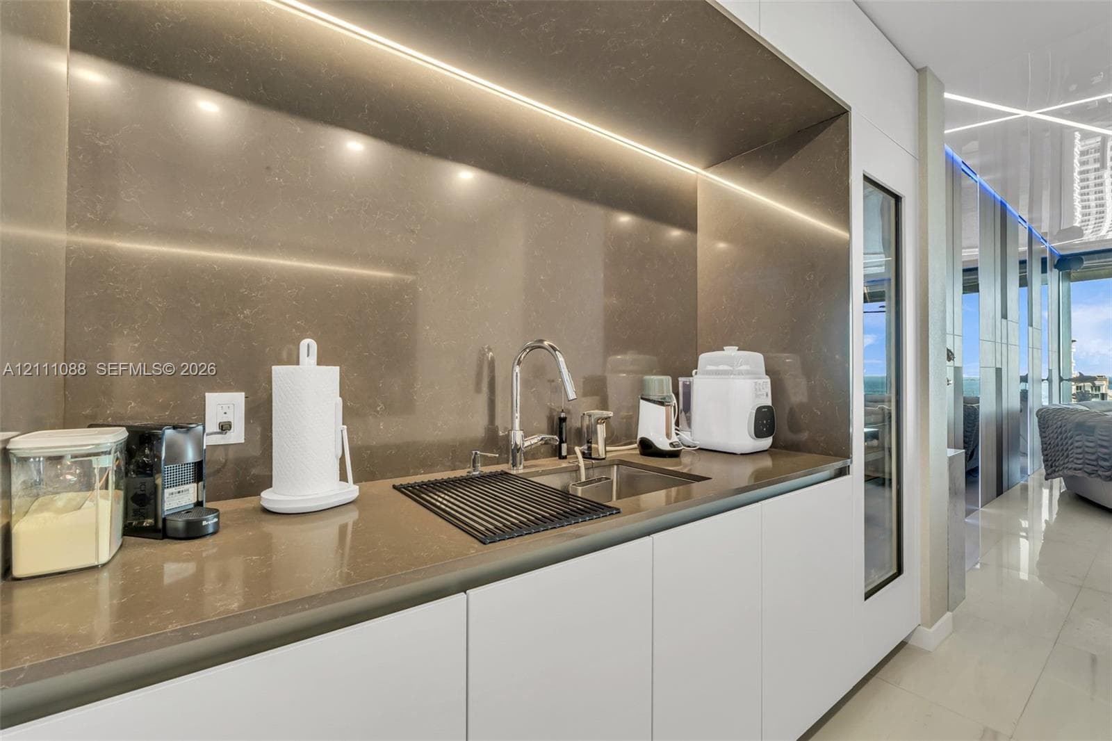 Sleek wet bar featuring minimalist flat-panel cabinetry, smooth stone-finish countertops, full-height backsplash, undermount sink with chrome gooseneck faucet, and integrated recessed LED under-cabinet lighting