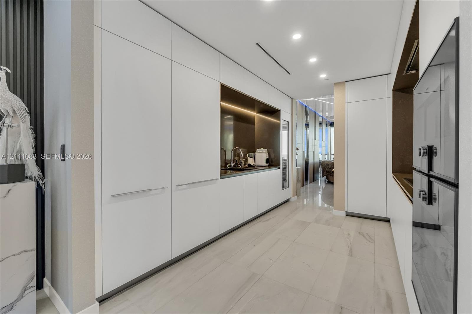 Sleek floor-to-ceiling white cabinetry, integrated recessed lighting, stone-finish tile flooring, built-in dual wall ovens, and a recessed metallic-finish backsplash