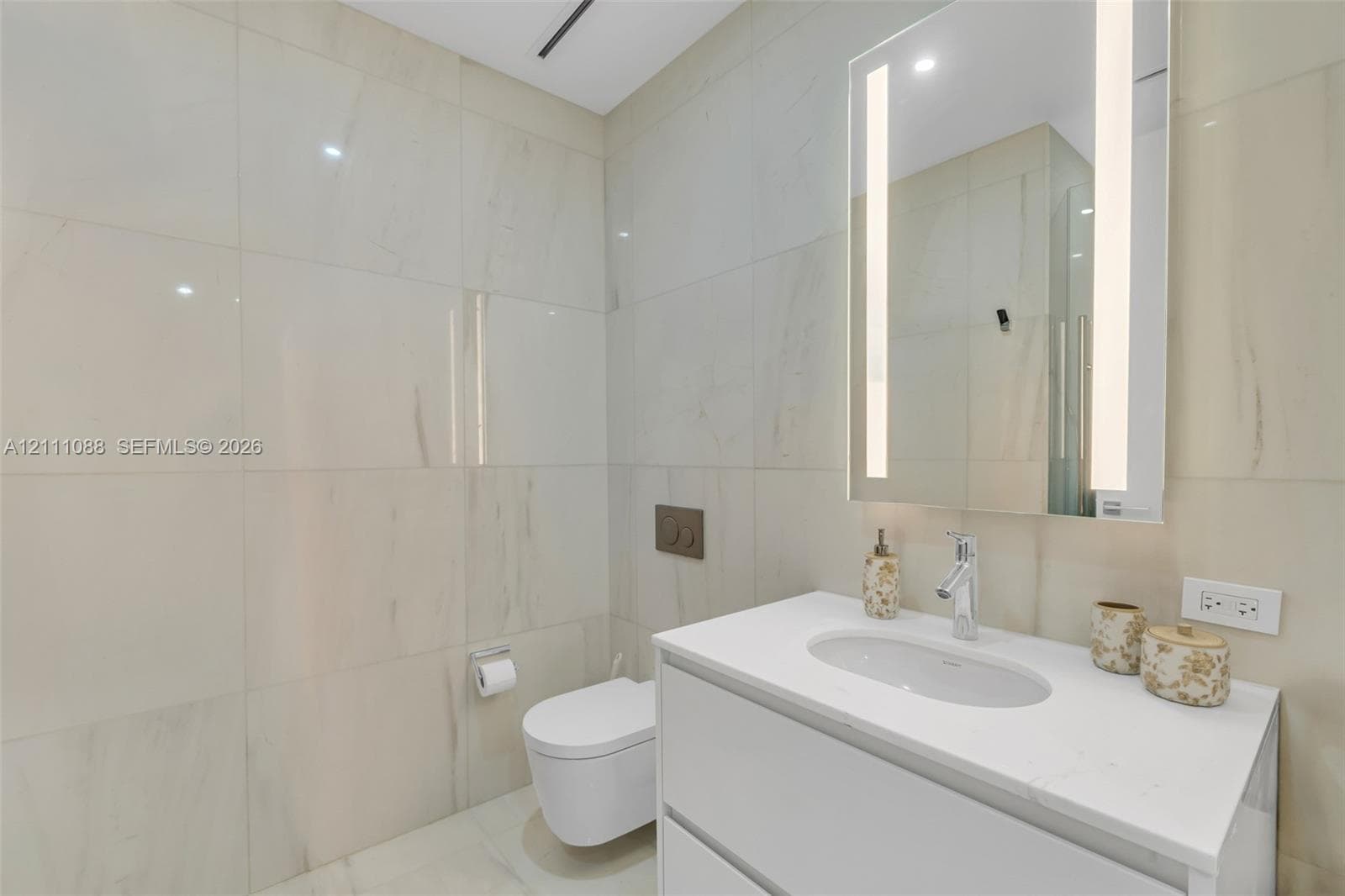 Full-height wall tiling, wall-mounted commode with a dual-flush actuator plate, floating vanity with a white countertop and integrated sink, and an LED-integrated vanity mirror