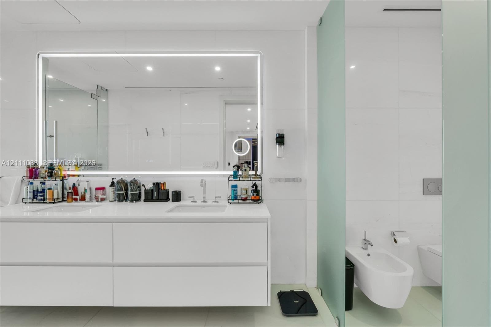 Floating white vanity with dual rectangular undermount sinks, large LED-backlit mirror, wall-to-wall stone-finish tiling, and wall-mounted bidet and toilet