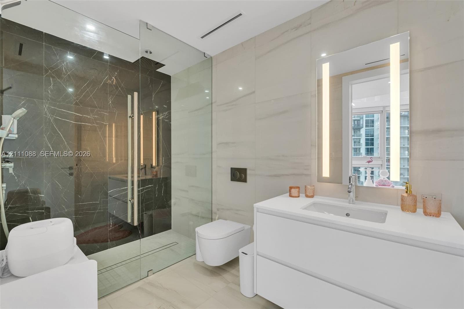 Bathroom featuring large-format light-toned wall tile, glass-enclosed shower with dark contrasting tile, wall-mounted toilet, white double-drawer vanity, and a rectangular mirror with integrated vertical lighting