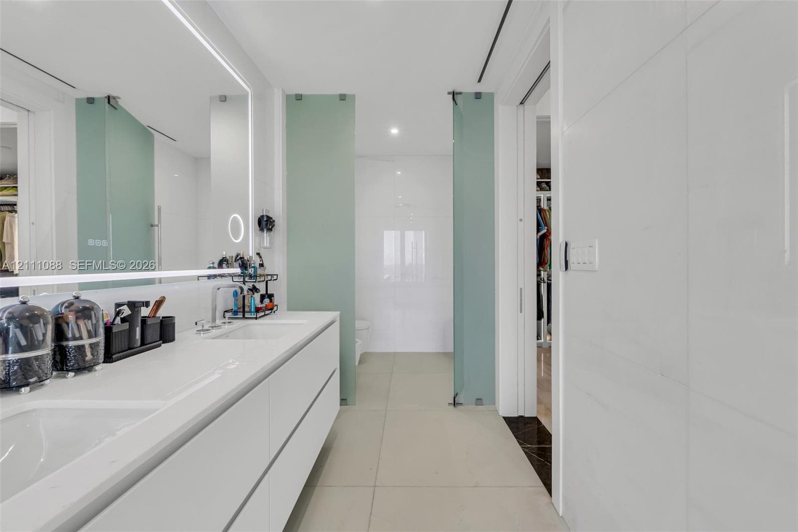 Minimalist dual-vanity featuring integrated sinks, quartz countertop, wall-to-wall backlighting, large-format tile flooring, and frosted glass privacy partitions
