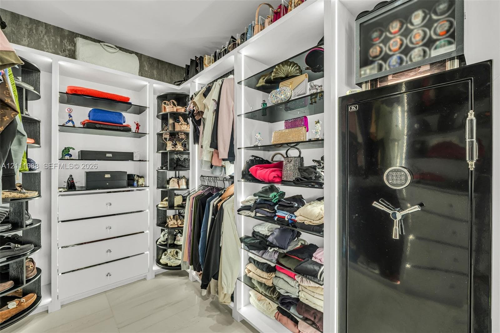 Custom closet system featuring white built-in cabinetry, vertical integrated LED lighting, open glass shelving, a rotating shoe rack, and an integrated heavy-duty floor safe