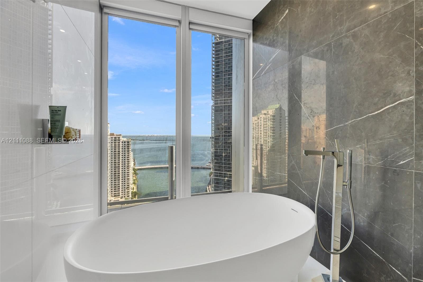 Freestanding soaking tub with polished chrome floor-mounted faucet, large-format grey stone wall tile, and expansive floor-to-ceiling windows