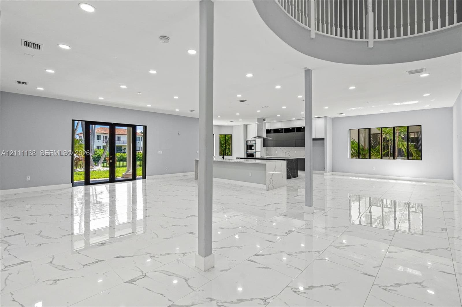 Open-concept interior featuring large-format stone-finish flooring, recessed lighting, a kitchen island with stone-finish countertop, structural support columns, and a mezzanine balcony with a metal railing
