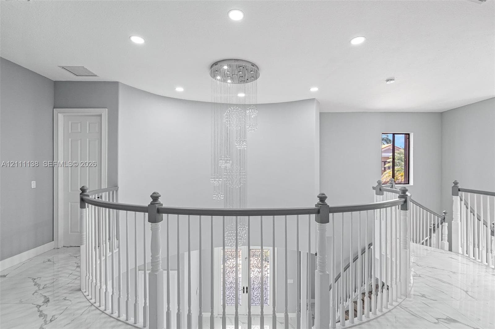 Curved architectural wall featuring a multi-tiered crystal chandelier, polished stone flooring, and a balustrade staircase with wood-finish handrails