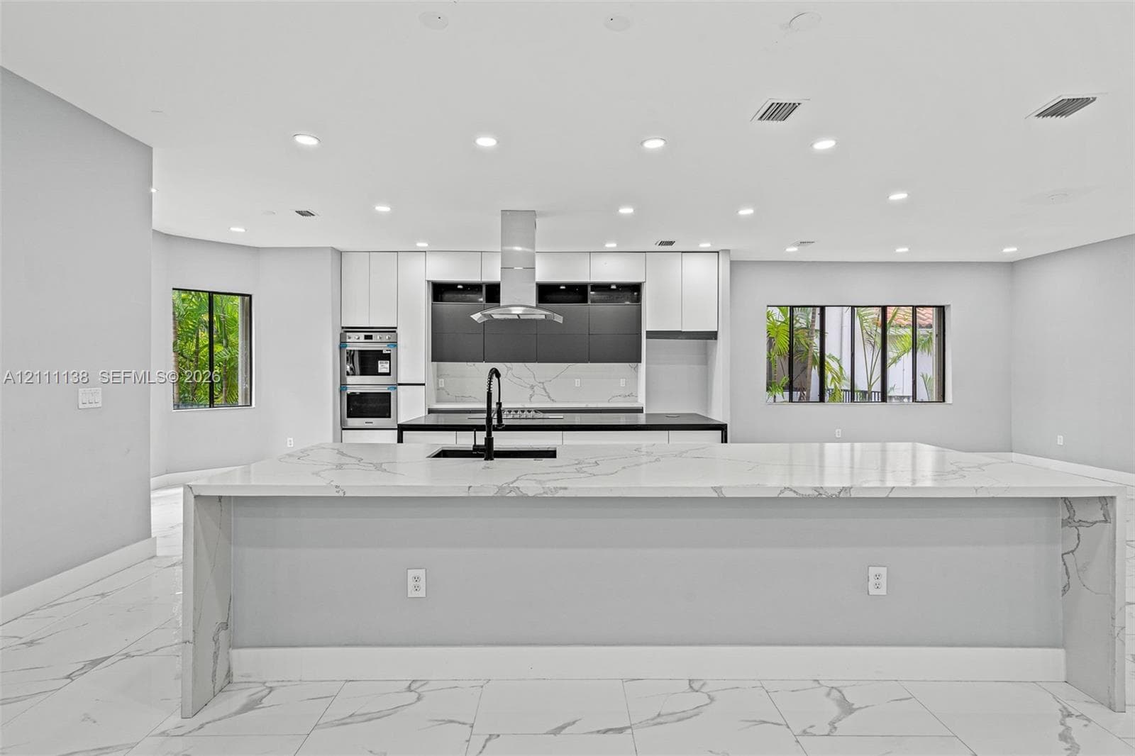 Kitchen featuring a large waterfall-edge island with stone-finish countertops, matte black faucet, integrated stainless steel double oven, two-tone cabinetry, and recessed lighting