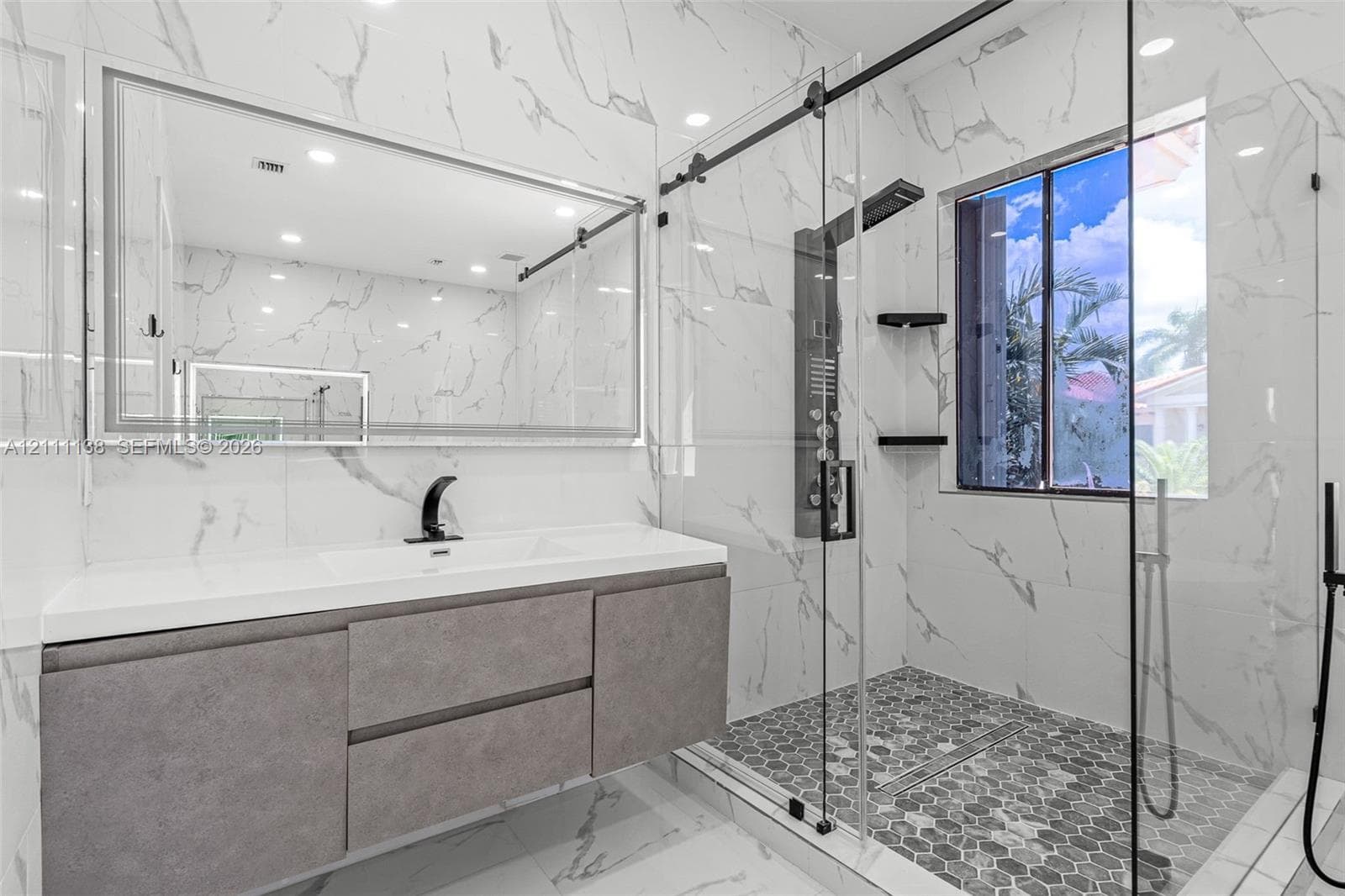 Floating vanity with integrated sink, floor-to-ceiling large-format tile walls, walk-in shower with glass enclosure, multi-head shower panel, and hexagonal tile flooring