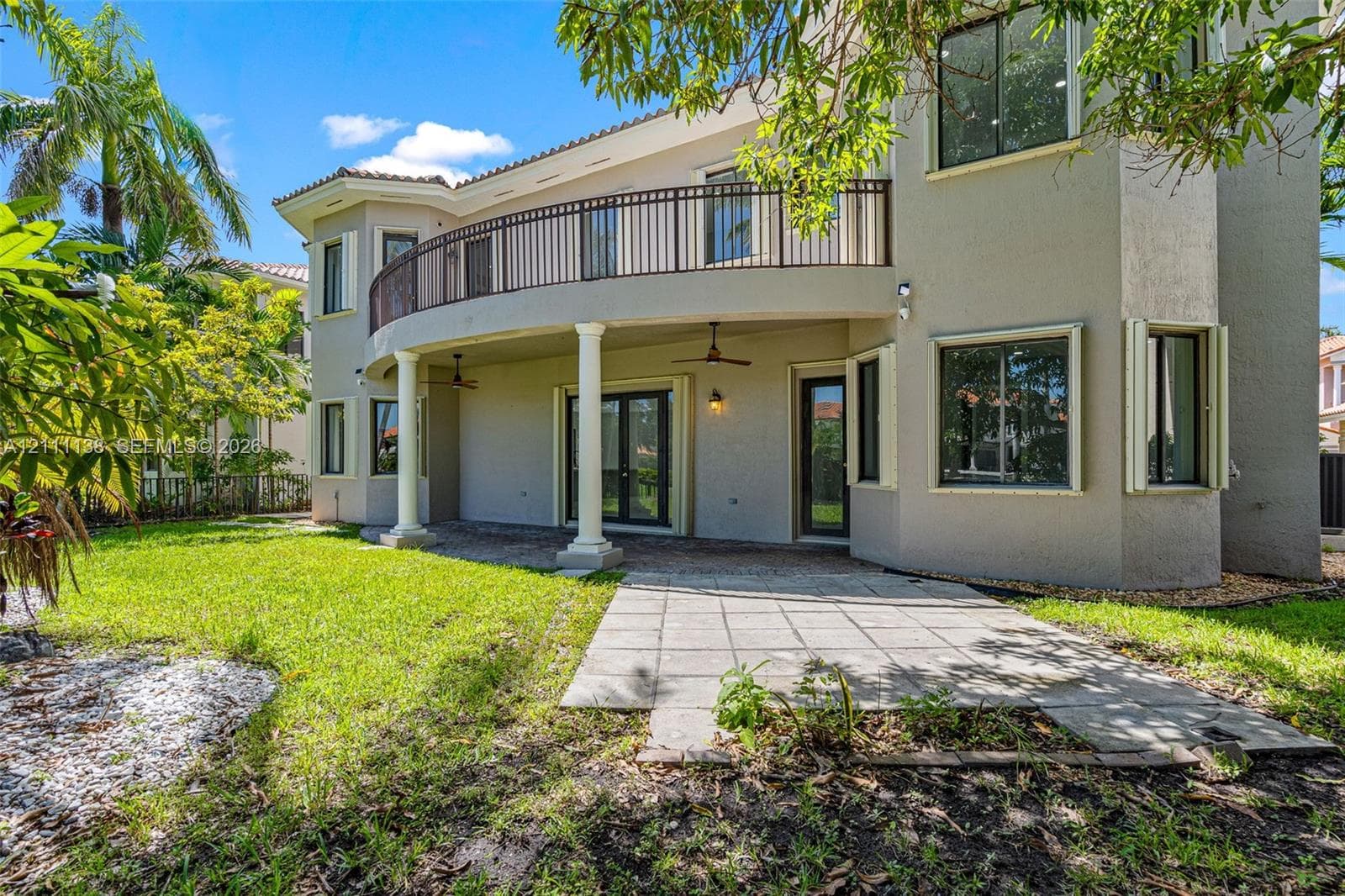 Stucco exterior featuring a covered patio, paved hardscaping, a second-story curved balcony with metal railing, and structural columns