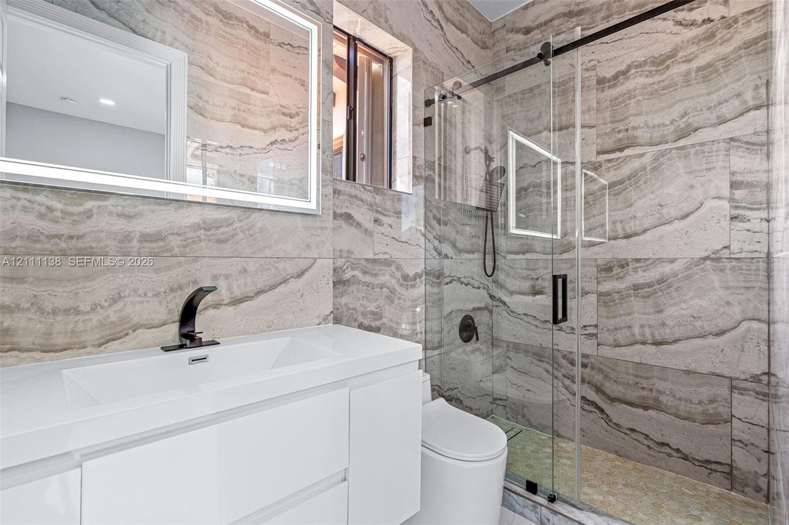 Modern vanity with a rectangular integrated sink, wall-mounted backlit mirror, walk-in shower with sliding glass door, and large-format natural stone wall tiling