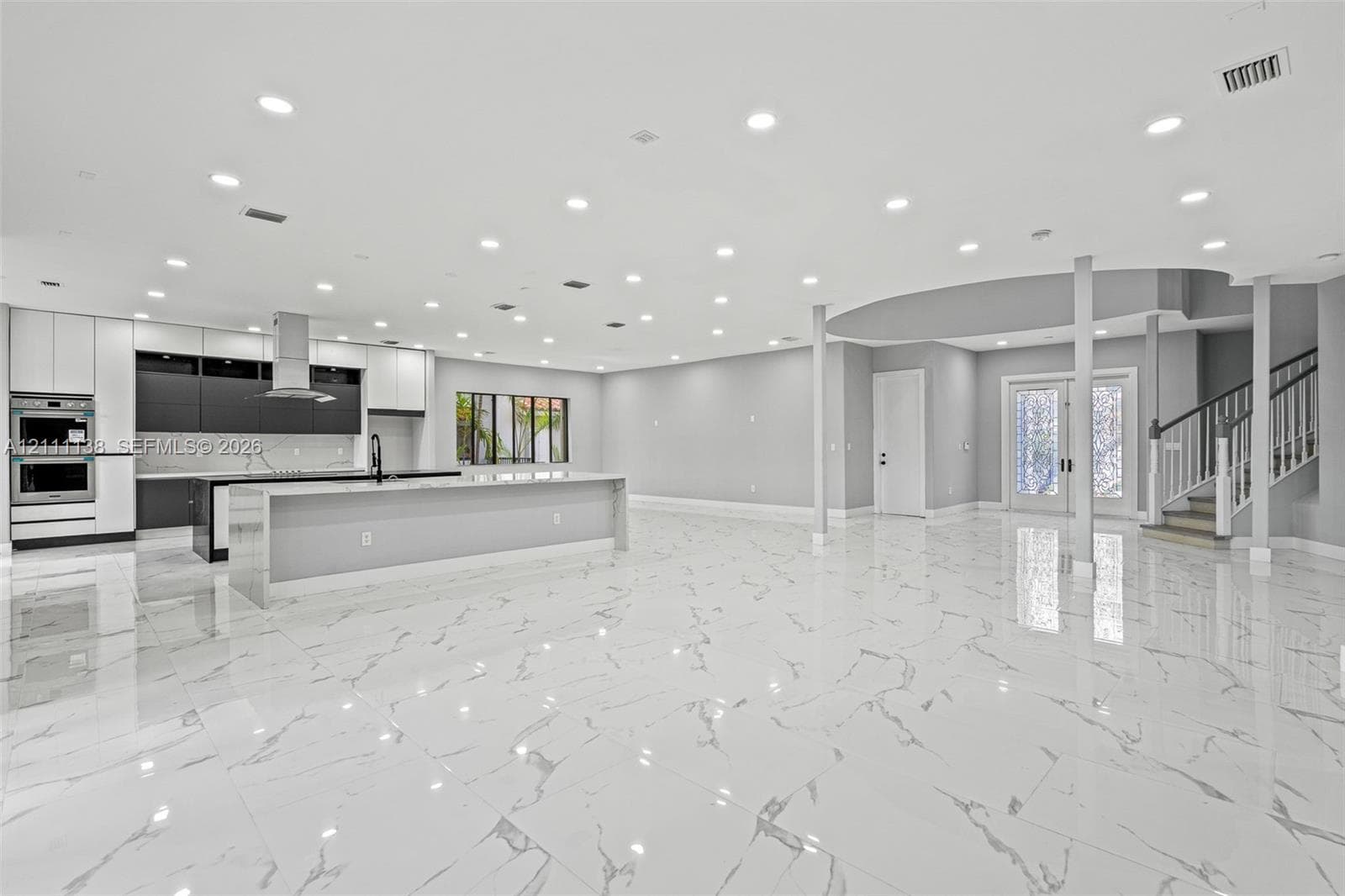 Open-concept interior featuring large-format tile flooring, recessed lighting, a modern kitchen with a dual-tone cabinetry finish, a central island, and a staircase with a wooden handrail