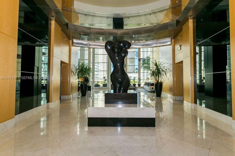 Grand foyer featuring light-toned stone flooring, full-height wall panels, curved glass mezzanine railing, and floor-to-ceiling windows