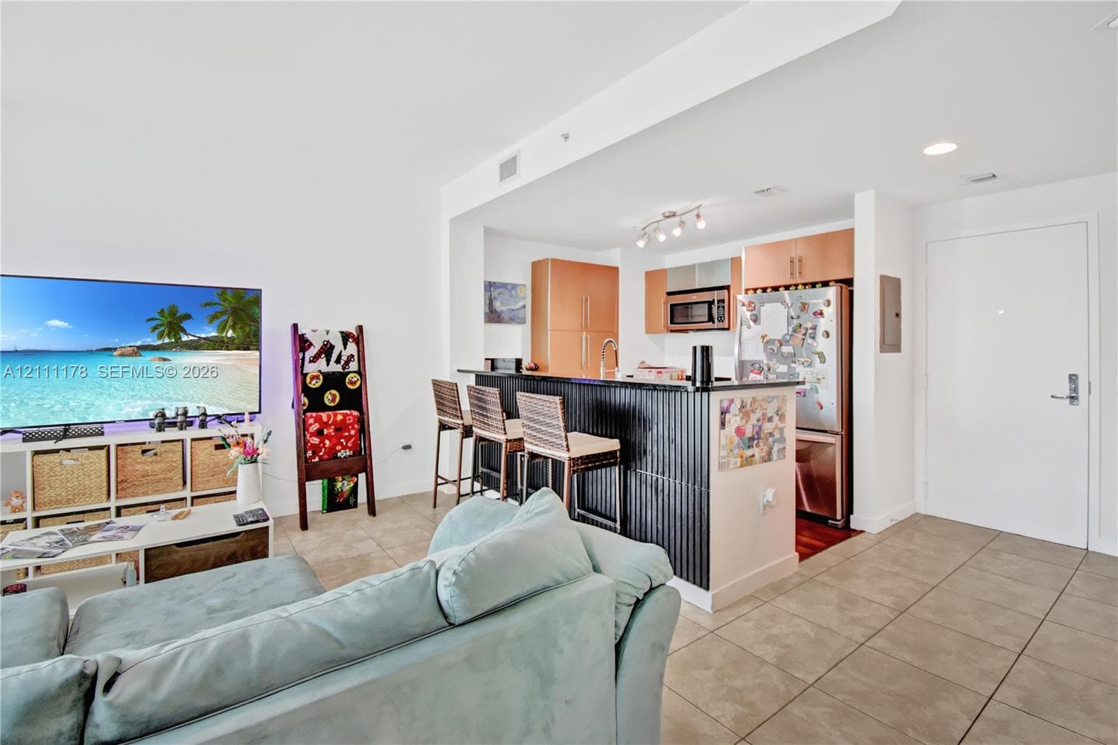 Open-concept kitchen featuring wood-finish cabinetry, stainless steel appliances, and a breakfast bar with stone-finish countertop and corrugated metallic base, all complemented by large-format tile flooring