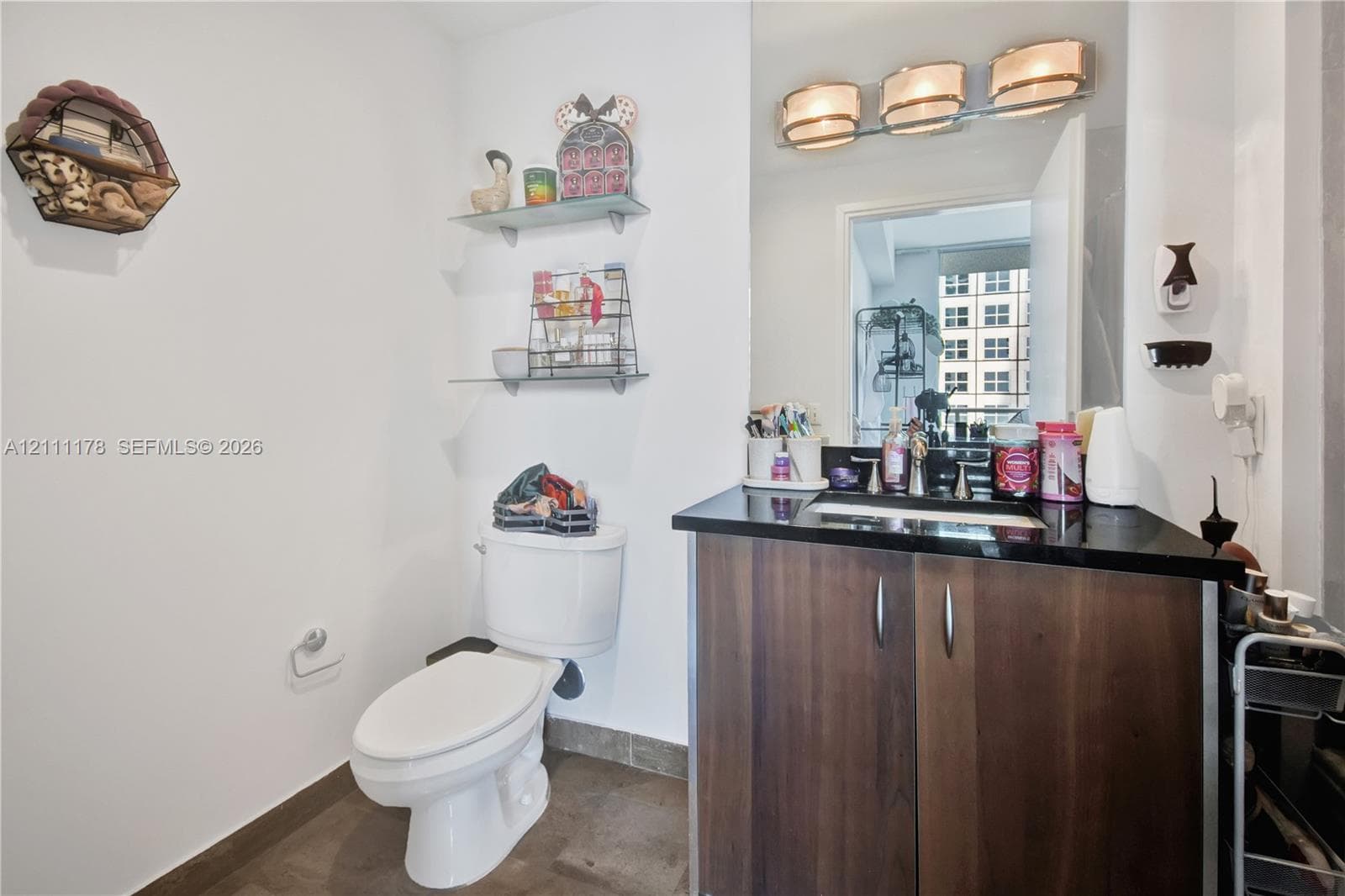 Vanity featuring a dark stone countertop, integrated sink, and wood-finish cabinetry, complemented by a three-light fixture, mirror, and glass wall shelving