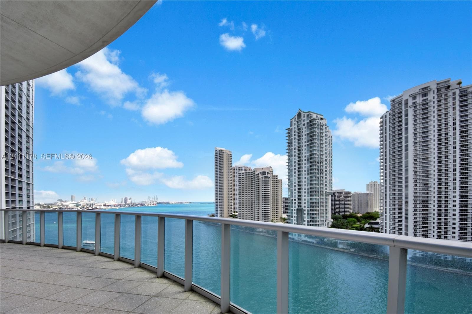 Covered balcony featuring concrete flooring and a glass-paneled railing system overlooking the water and metropolitan skyline