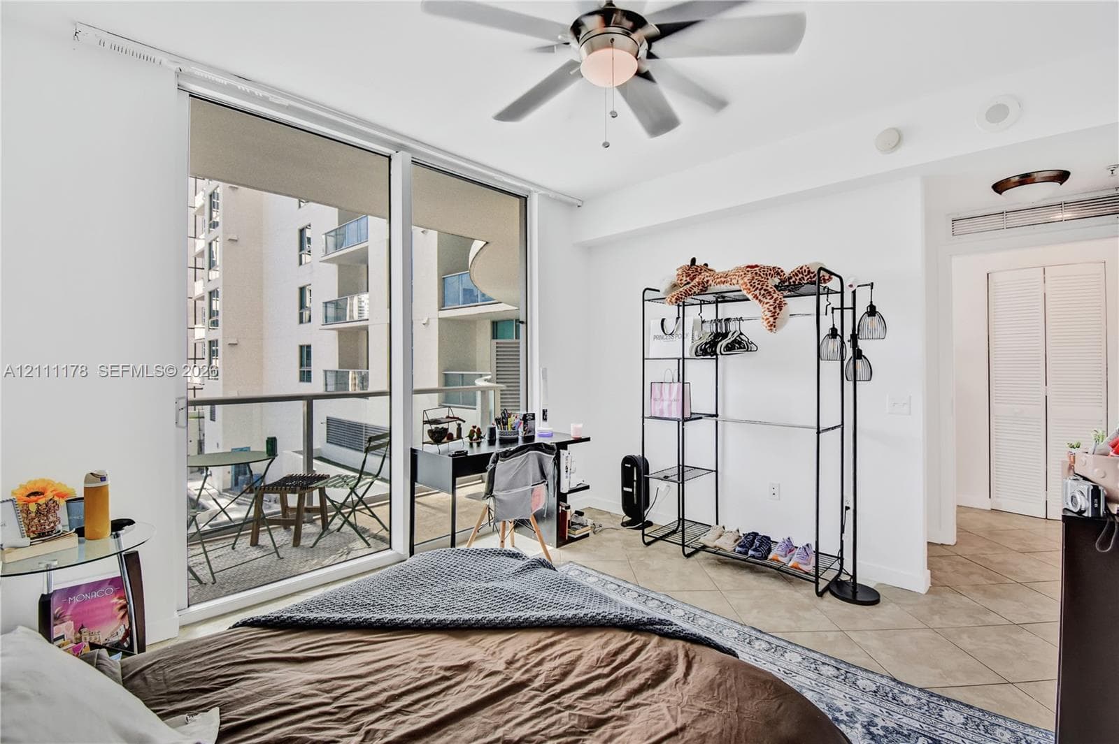 Floor-to-ceiling glass sliding doors, private balcony with metal railing, ceiling fan with integrated lighting, neutral tile flooring, and louvered closet doors