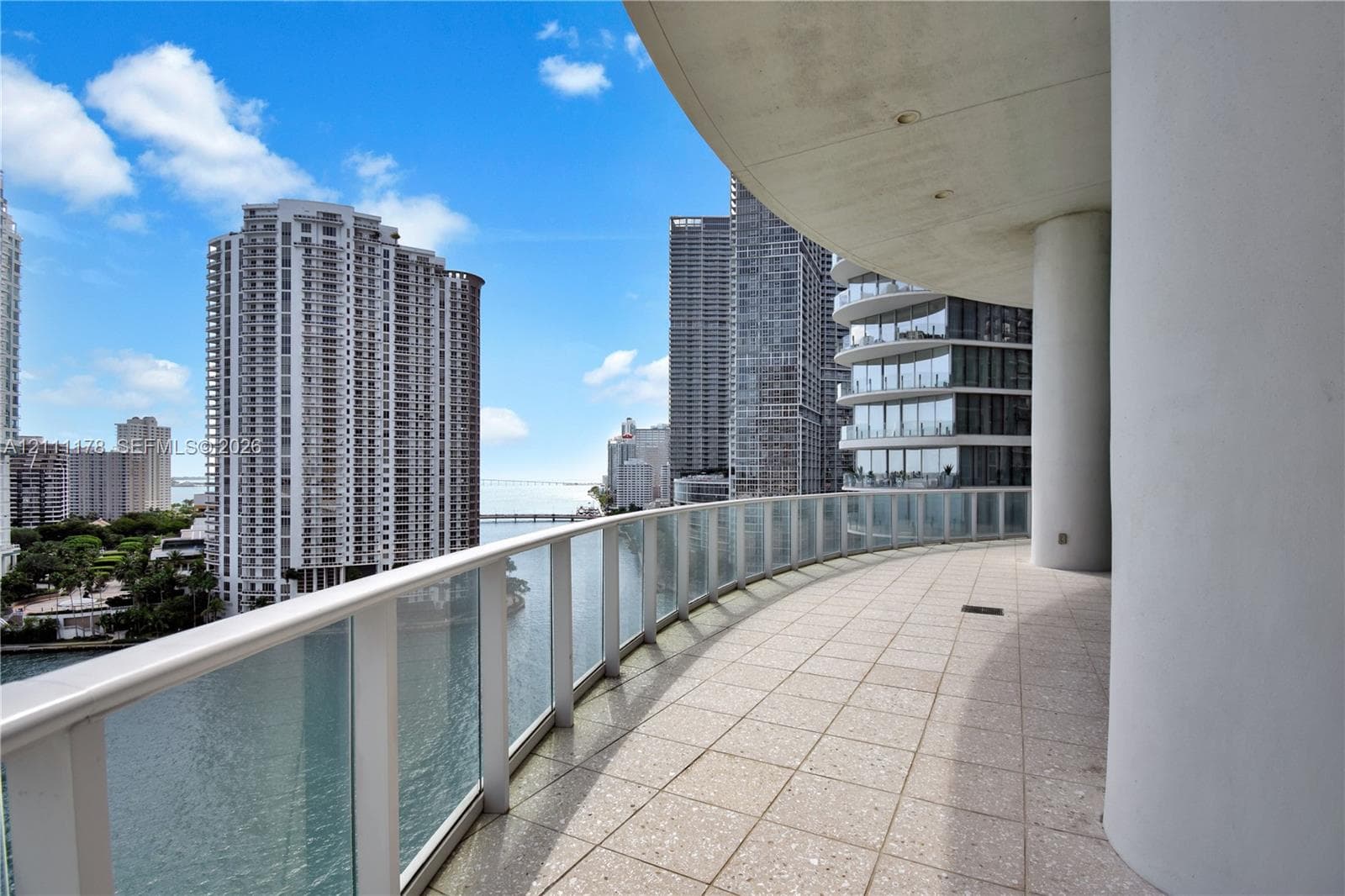 Expansive covered balcony featuring stone tile flooring, a glass-panel railing, and cylindrical structural columns
