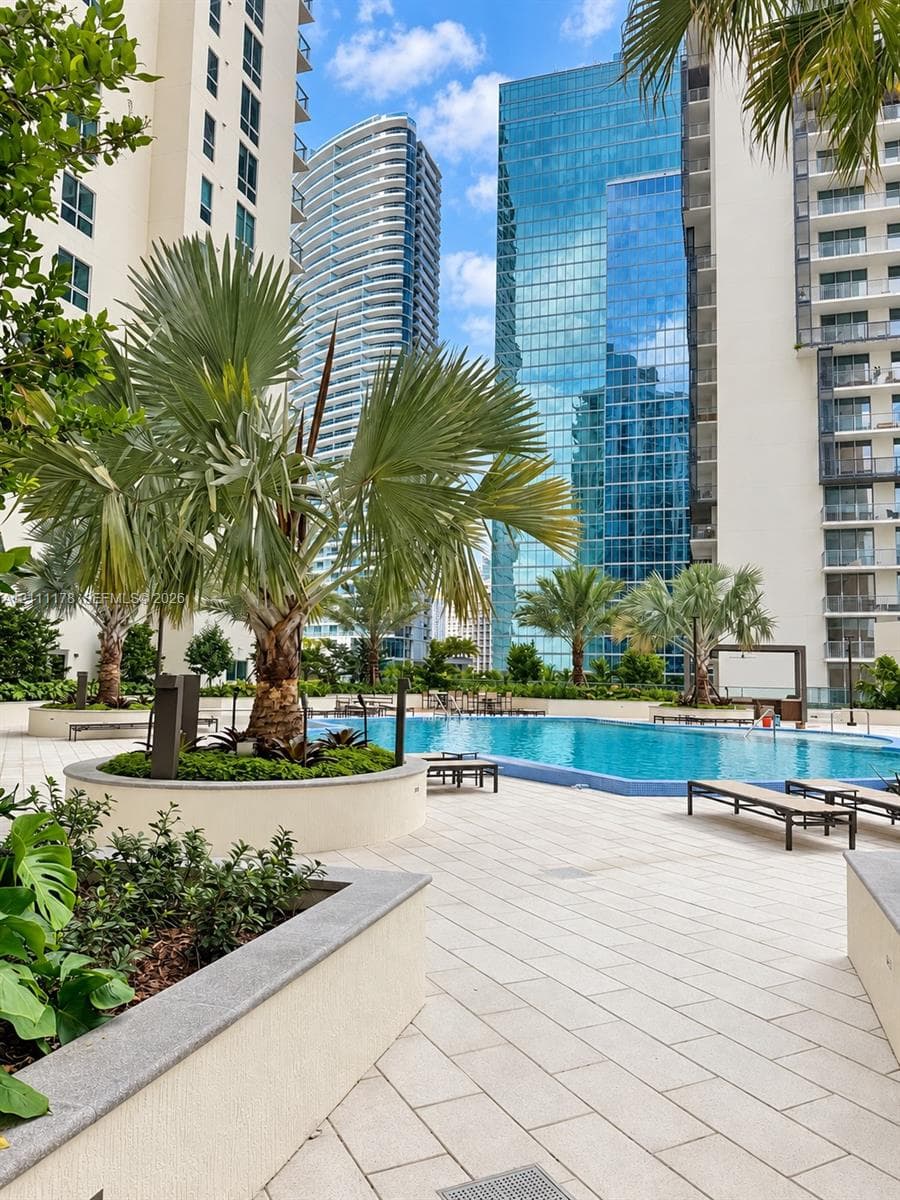 Swimming pool featuring light-toned paved decking, raised concrete planters, and mature palm trees surrounded by high-rise architecture