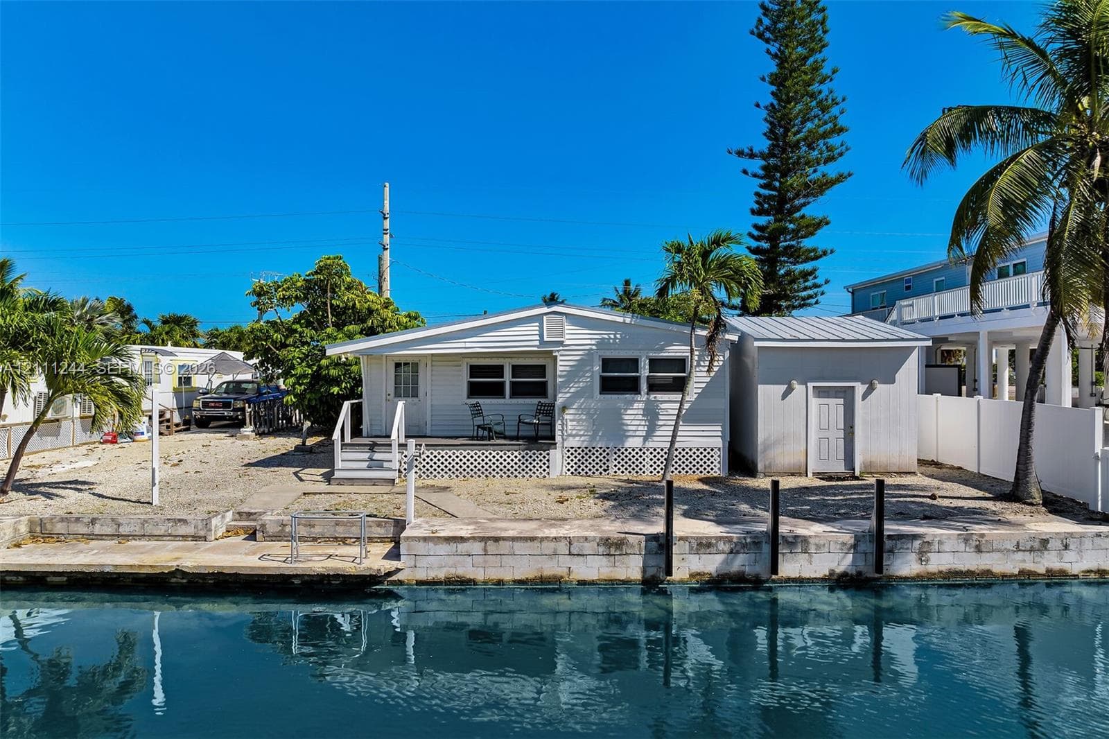 Waterfront exterior featuring light-colored horizontal siding, a raised deck with lattice skirting, an attached utility structure, and concrete seawall bulkhead