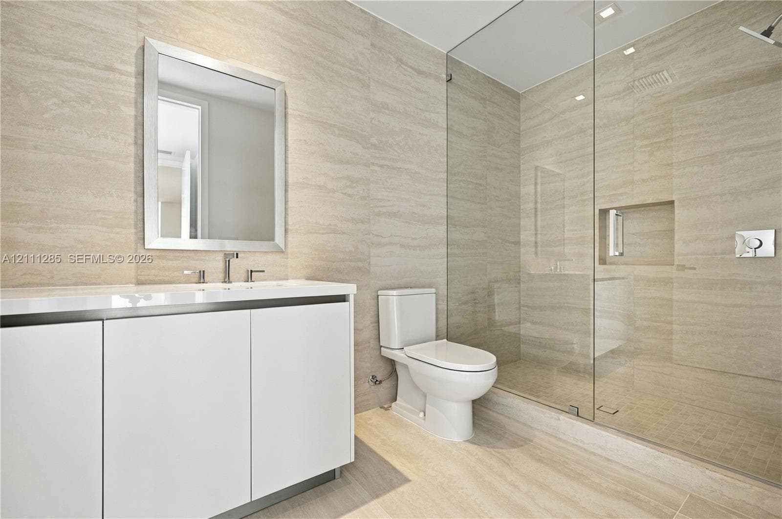 Wall-to-wall stone tile surfacing, walk-in glass-enclosed shower with a recessed shelf, white vanity with a rectangular sink, wall-mounted mirror, and a two-piece toilet