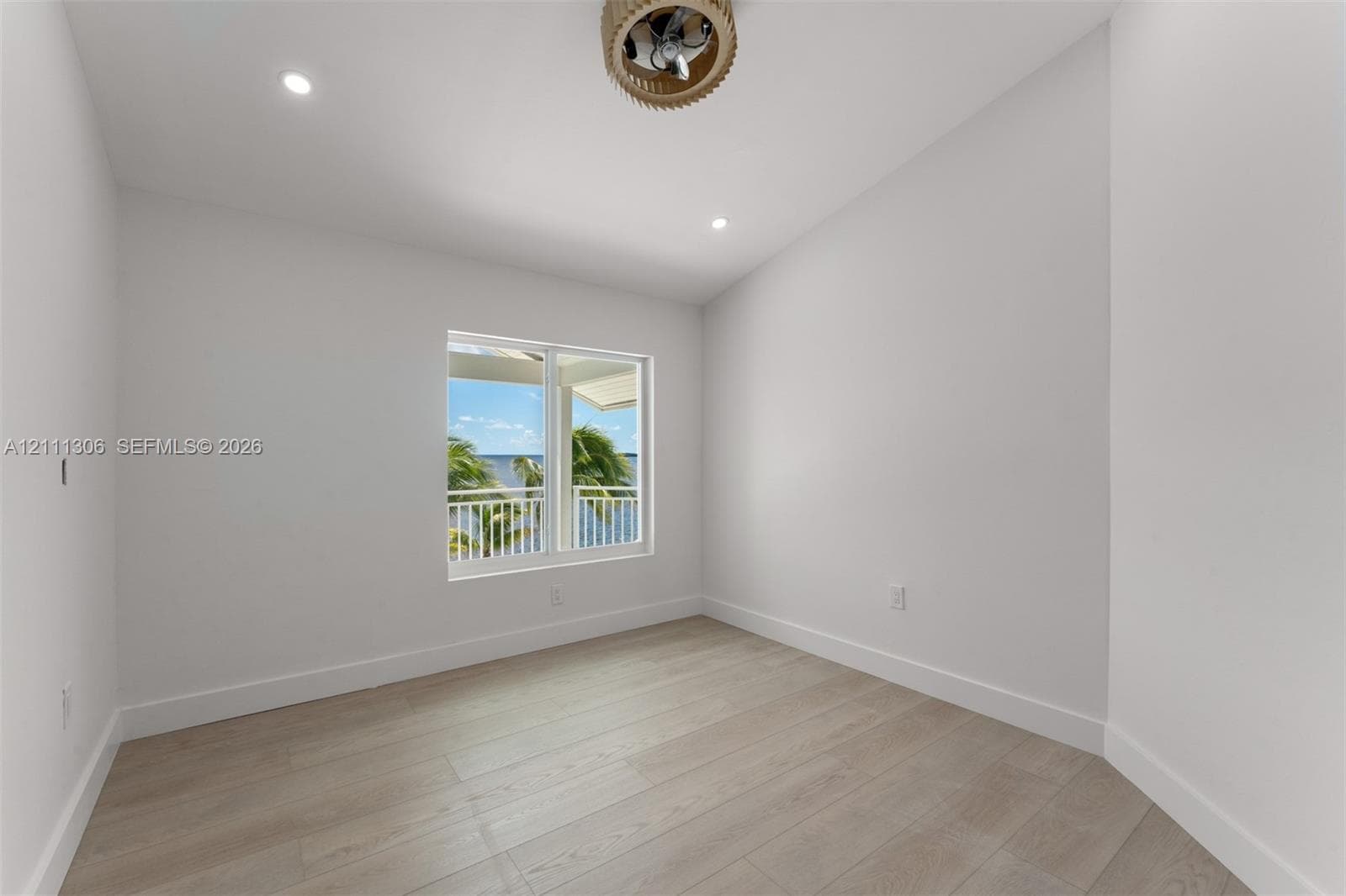 Neutral interior space featuring wood-finish flooring, recessed lighting, and a drum-style ceiling fan, with a window framing water and palm tree scenery