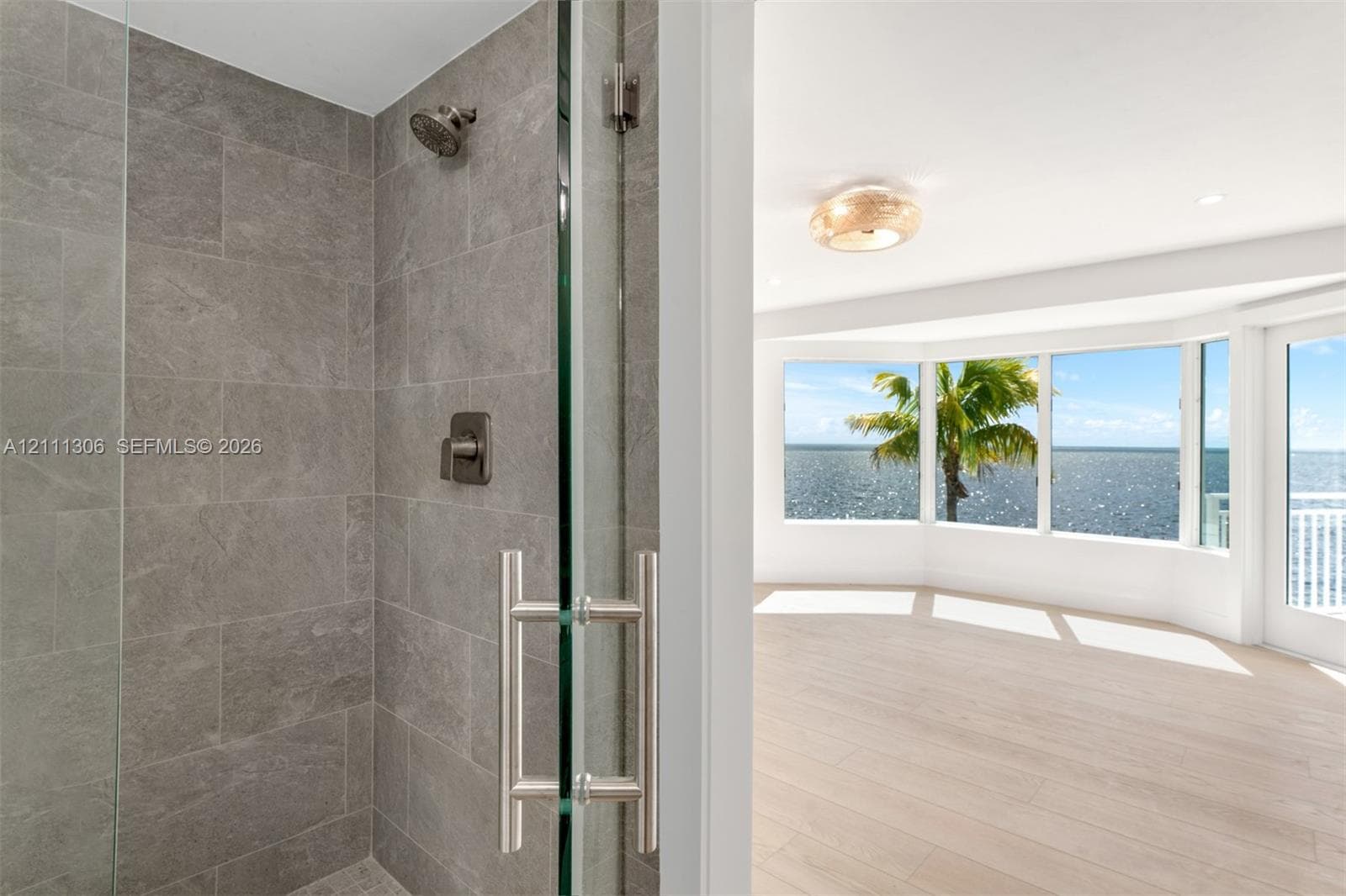 Shower enclosure featuring grey stone-finish tile, glass door with metallic hardware, and wall-mounted shower fixture, complemented by wood-finish flooring and bay windows featuring water surroundings