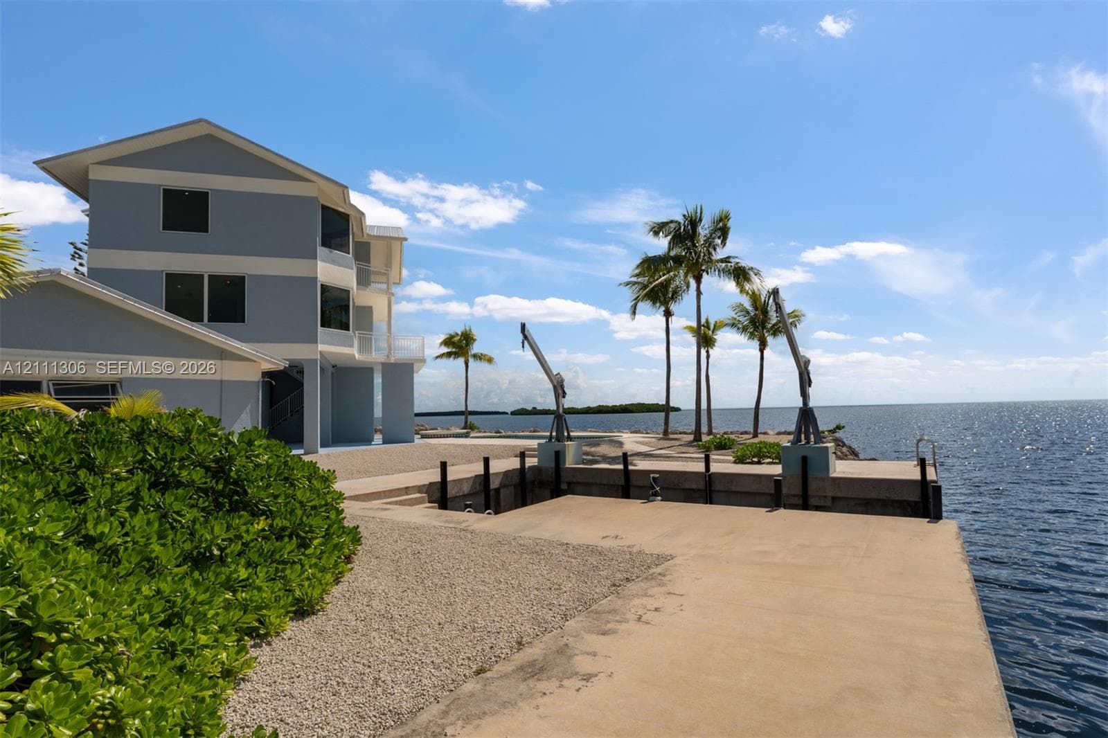 Three-story waterfront residence featuring a concrete seawall, dual boat davits, gravel landscaping, and mature palm trees
