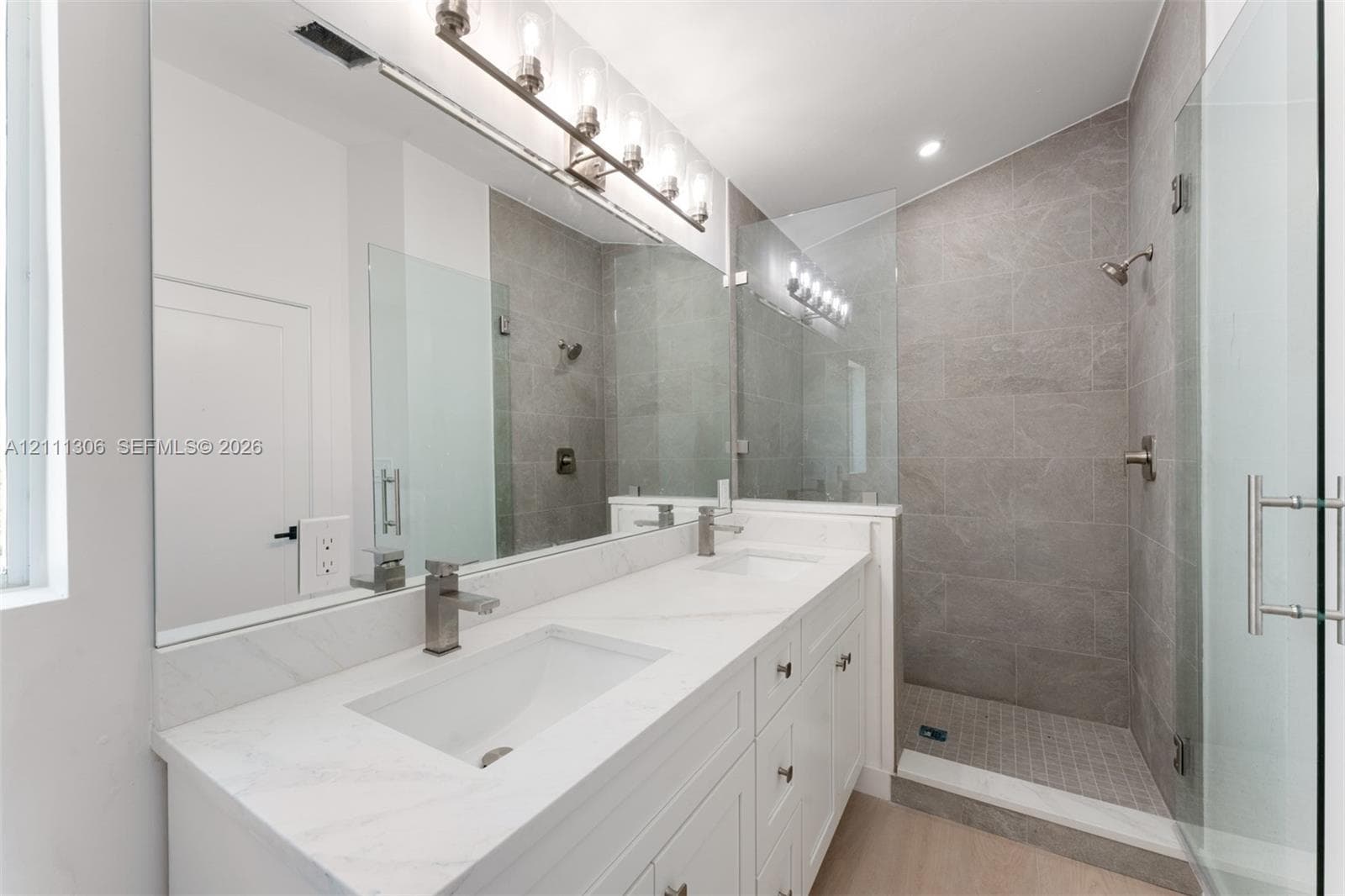 Double-vanity with stone countertop, walk-in shower featuring tile surround and glass enclosure, large wall-mounted mirror, and multi-bulb vanity lighting