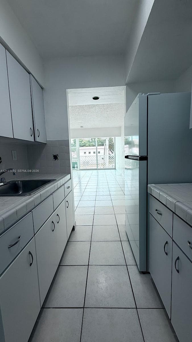 Galley-style kitchen featuring white cabinetry, tile countertops, stainless steel sink, and ceramic tile flooring