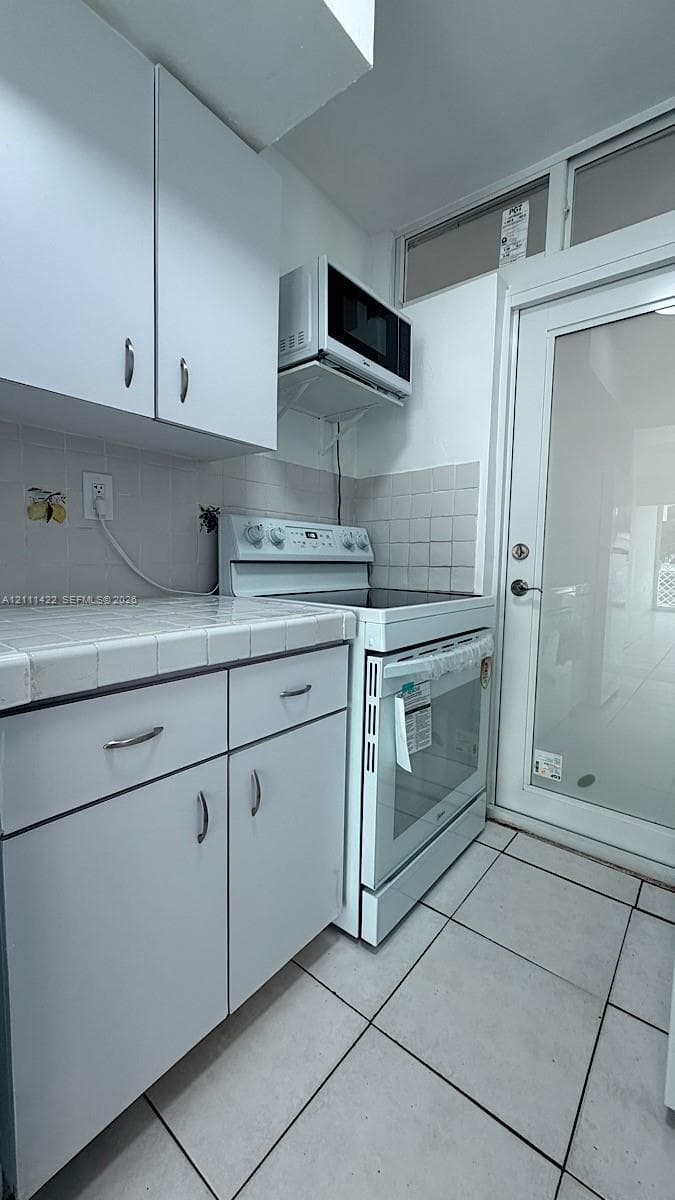 Kitchen featuring white cabinetry, ceramic tile countertops, light-colored tile flooring, integrated electric range, and a full-lite glass door