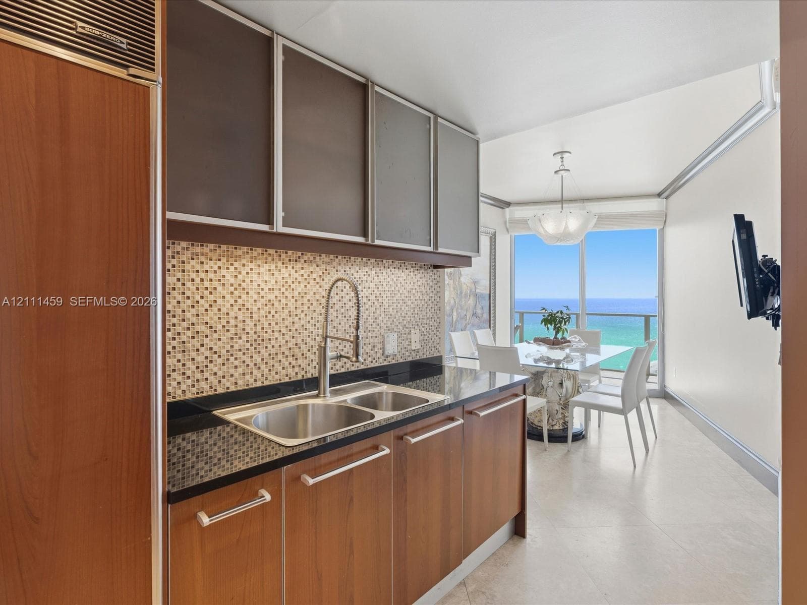 Kitchen featuring wood-finish cabinetry, a mosaic tile backsplash, dark solid-surface countertops, an integrated stainless steel refrigerator, and large-format floor tiling