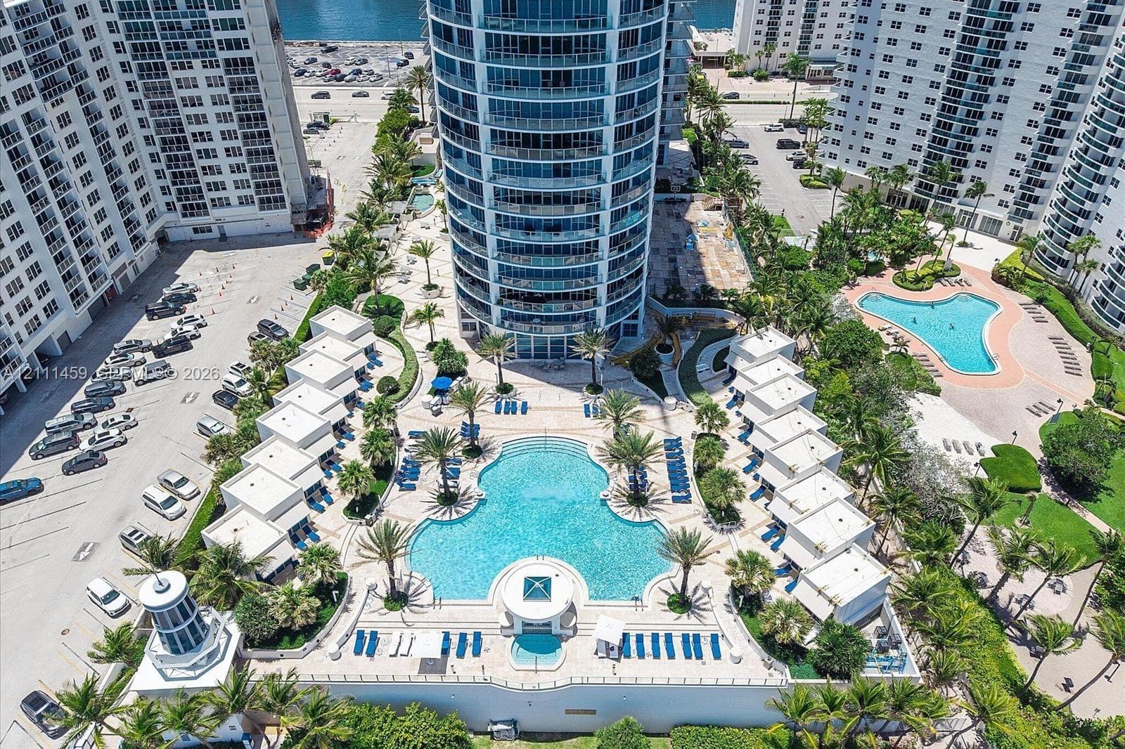 High-rise residential tower featuring a multi-level pool deck, multiple cabanas, palm tree landscaping, and paved hardscaping