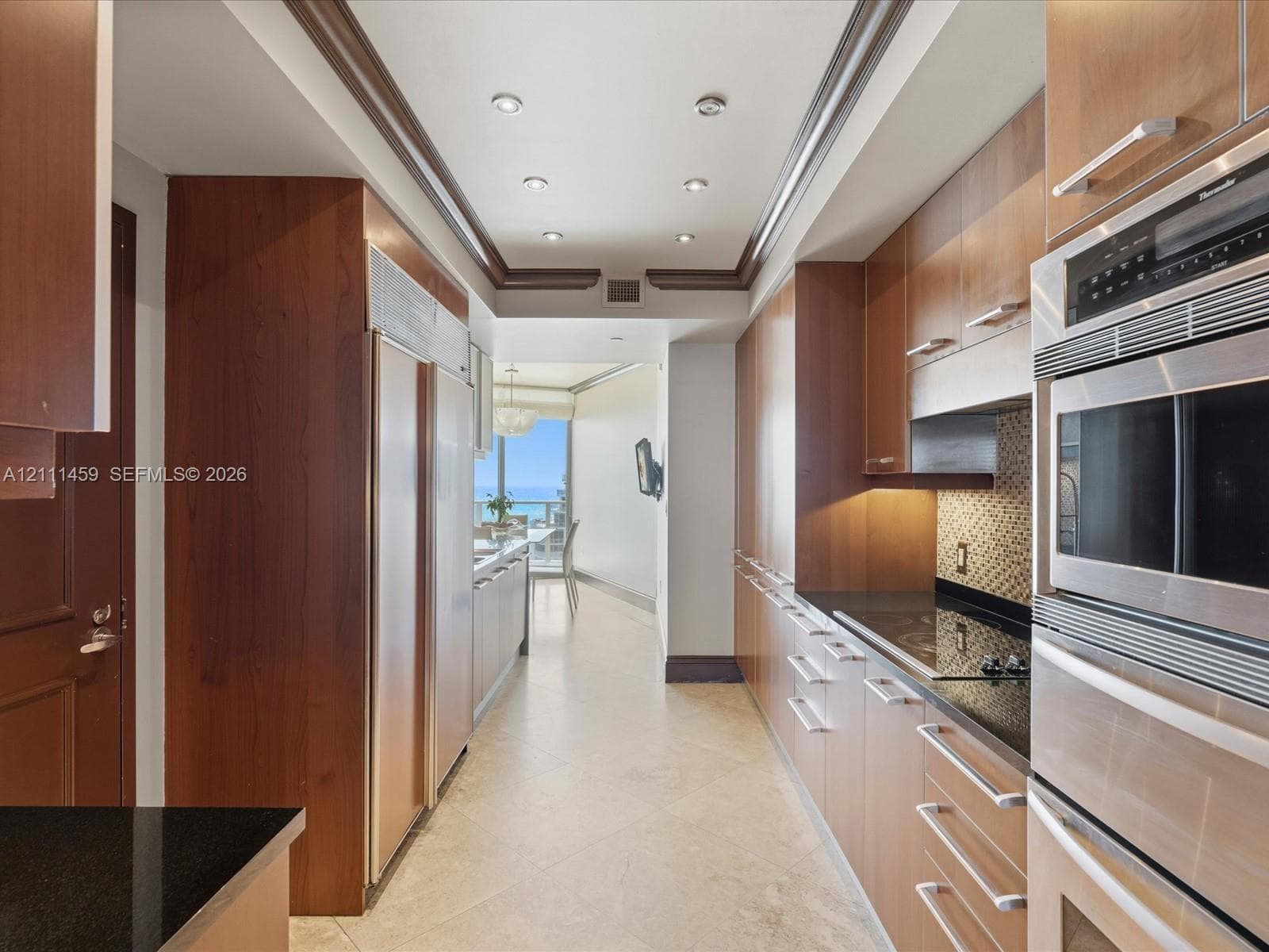 Galley kitchen featuring custom flat-panel wood cabinetry, black stone countertops, integrated stainless steel refrigeration, built-in double ovens, and recessed ceiling lighting